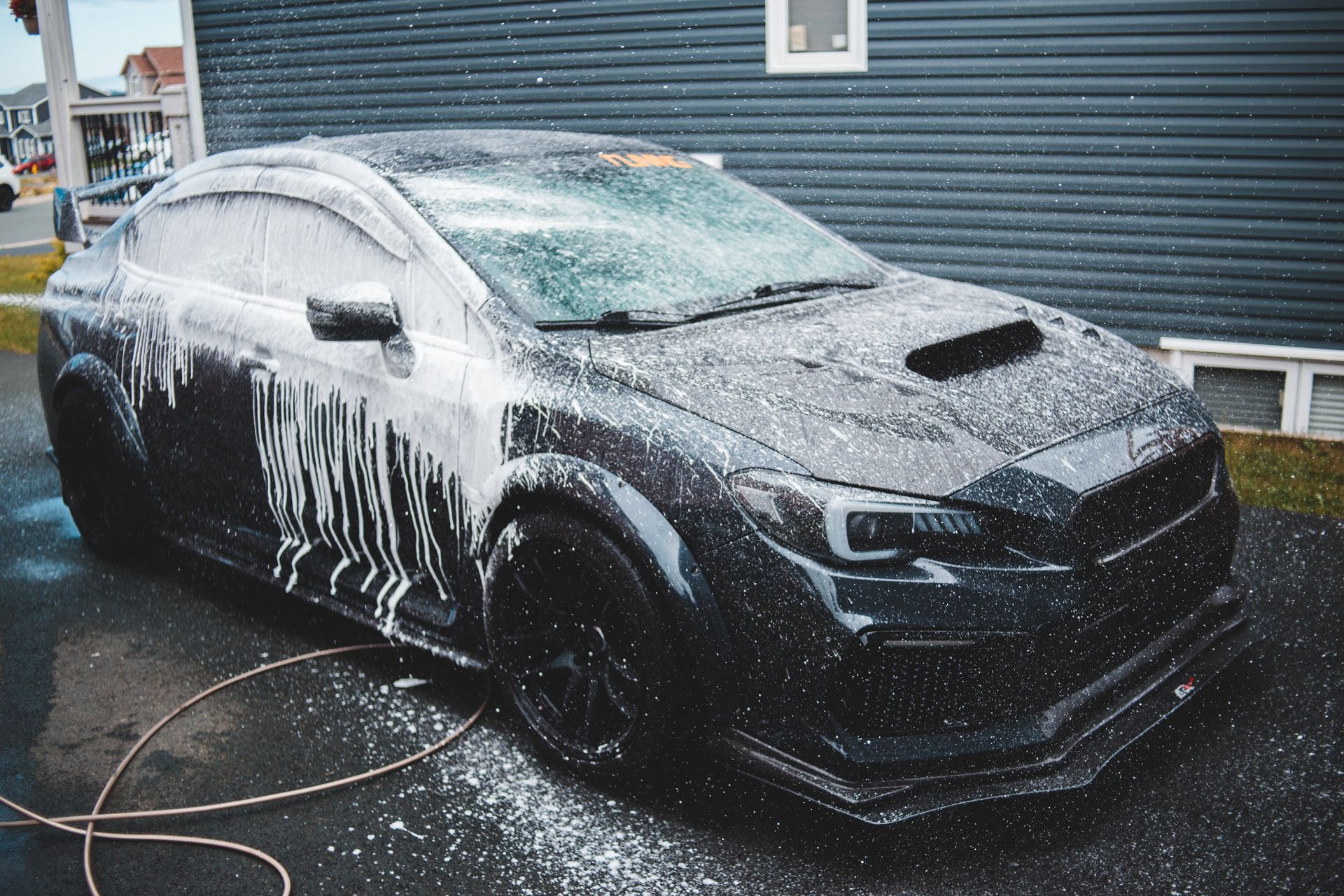 Black sedan being washed with foam outside a garage, with a hose on the wet ground