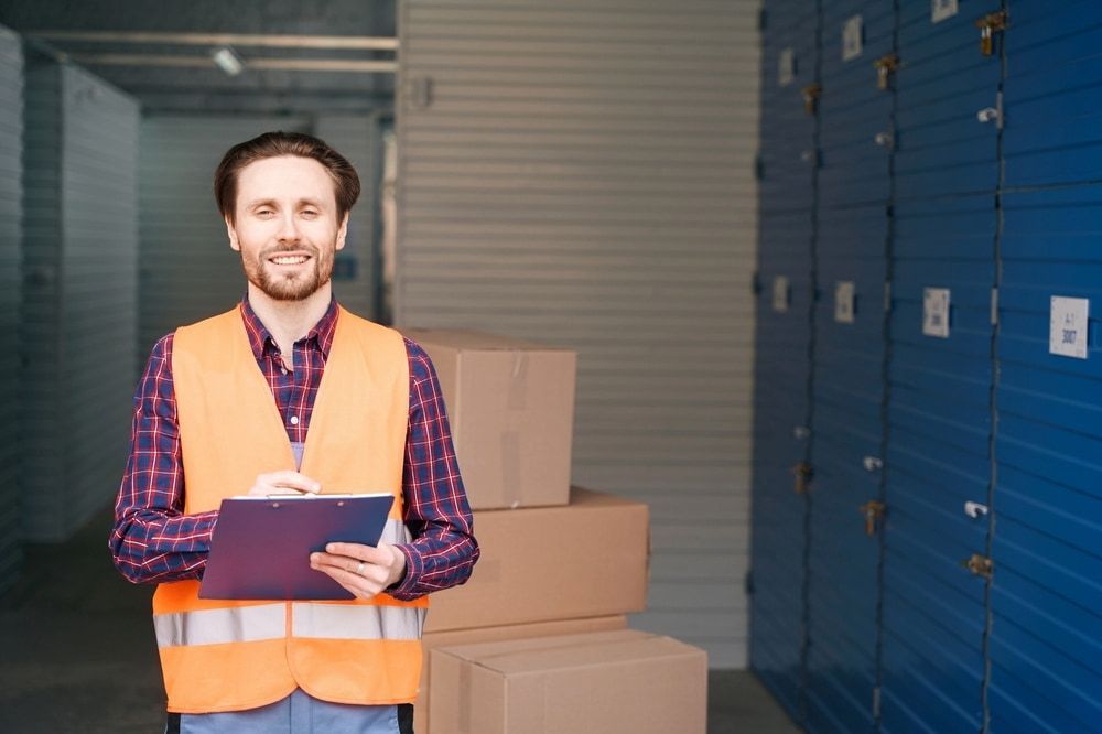 A Man In An Orange Vest Is Holding A Clipboard In A Warehouse — Newcastle Moving & Storage in Salt Ash, NSW