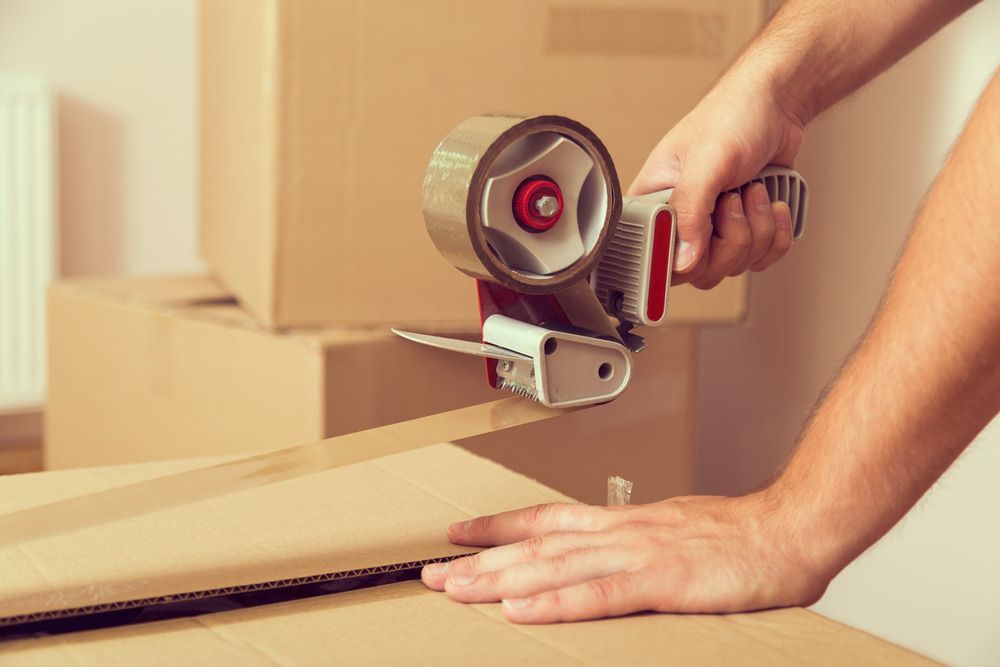 A Person Is Using A Tape Dispenser To Seal A Cardboard Box — Newcastle Moving & Storage in Cessnock, NSW