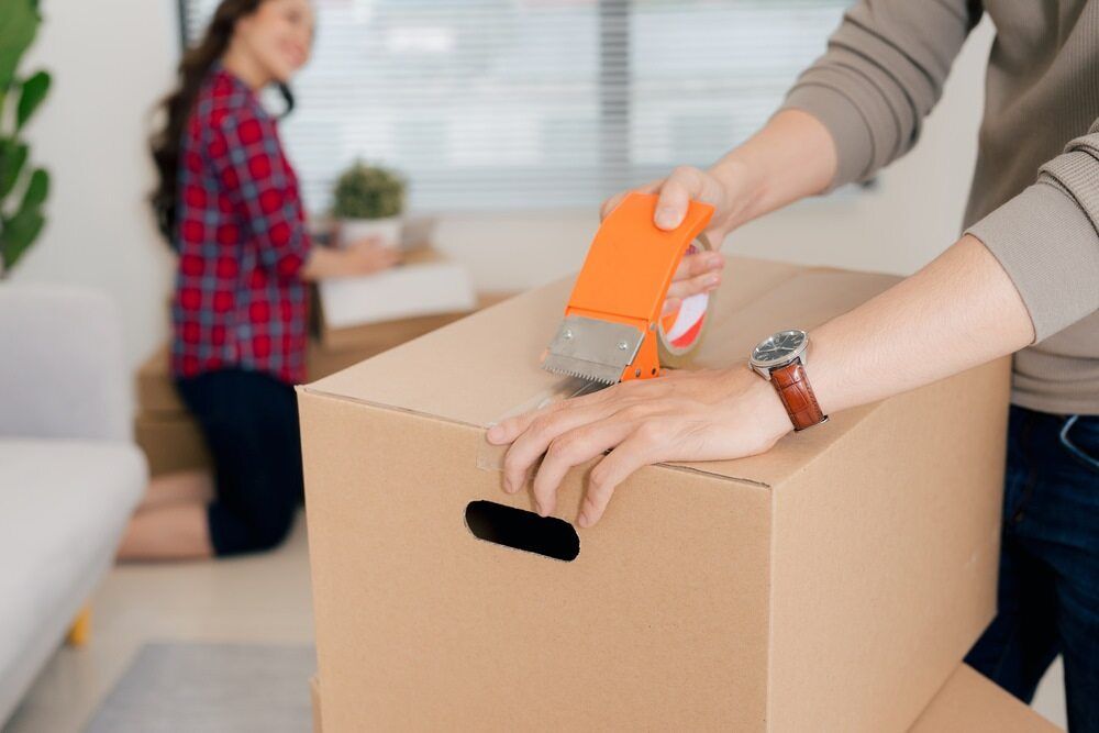 A Man Is Taping A Cardboard Box With Tape — Newcastle Moving & Storage in Swansea, NSW