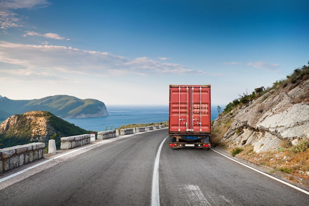 A Red Truck Is Driving Down A Mountain Road Next To The Ocean — Newcastle Moving & Storage in Salt Ash, NSW