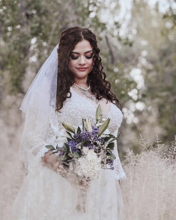 Bride in white lace gown holding a purple-and-white bouquet outdoors