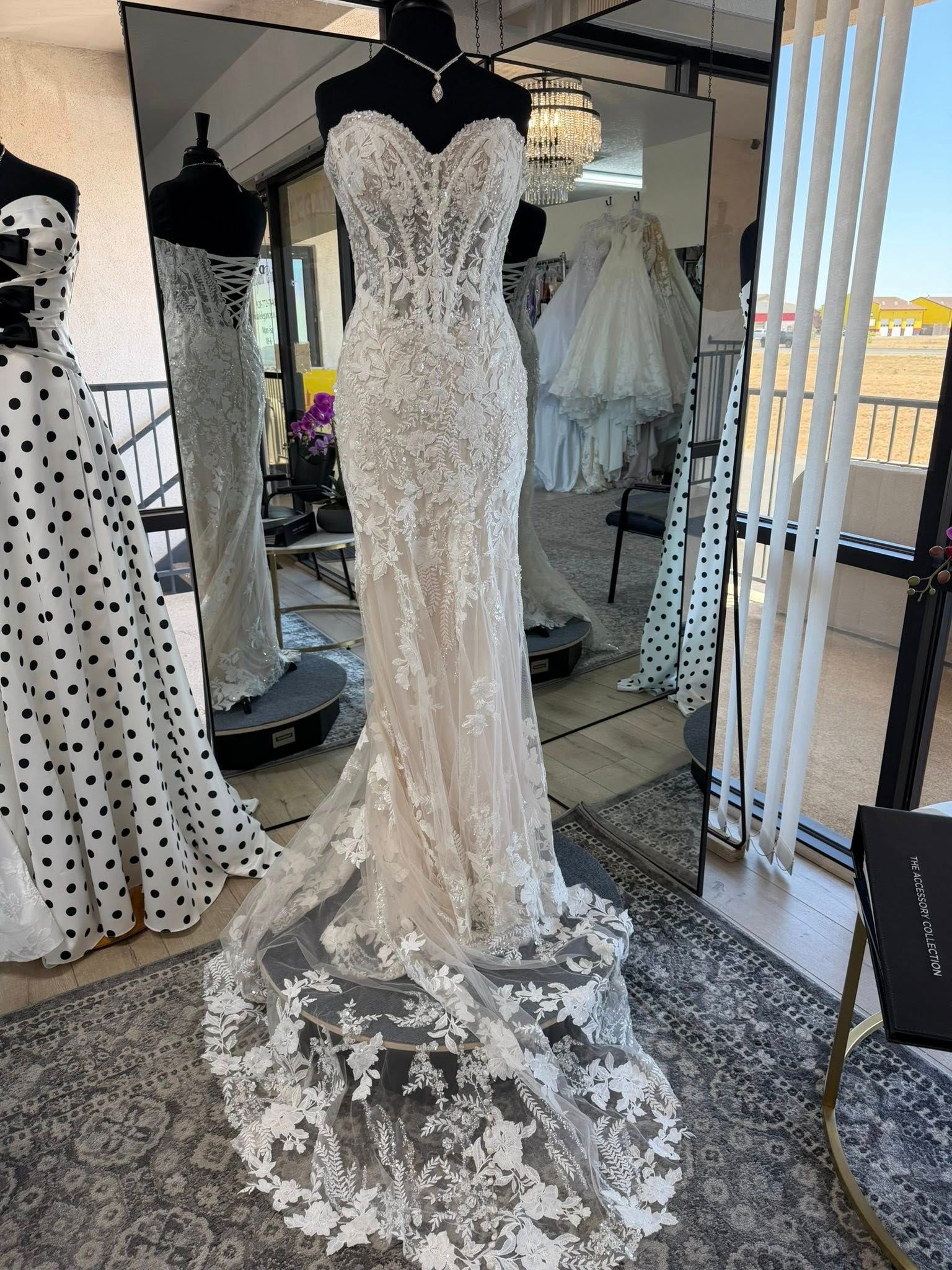White lace wedding dress on a mannequin in a boutique, with a long lace train and mirrored background.