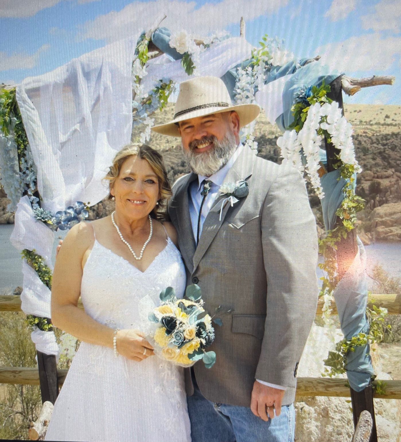 Couple in wedding attire posing outdoors by a white floral arch, holding a bouquet.