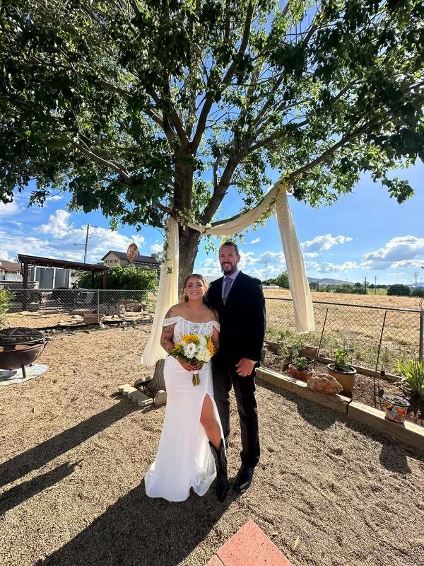 Bride and groom standing under a tree at an outdoor wedding, bride holding a bouquet and both smiling