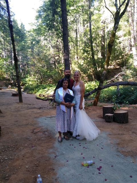 Two women in white dresses pose together on a wooded path with flowers scattered on the ground.