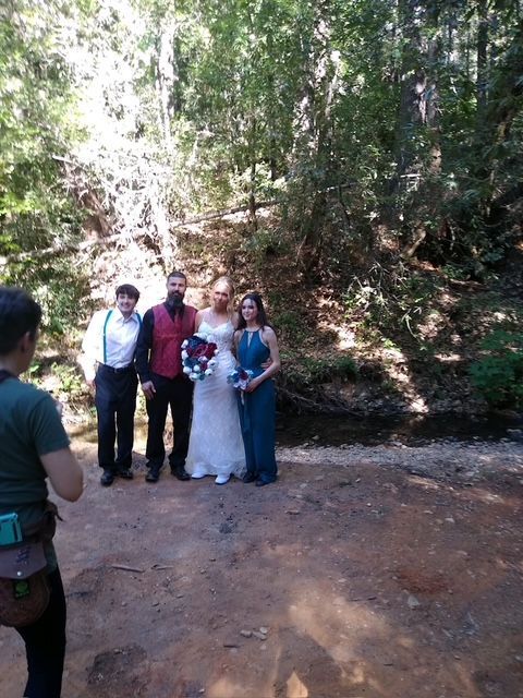 Wedding party posing outdoors in a wooded clearing, with a bride in white and others in formal attire.