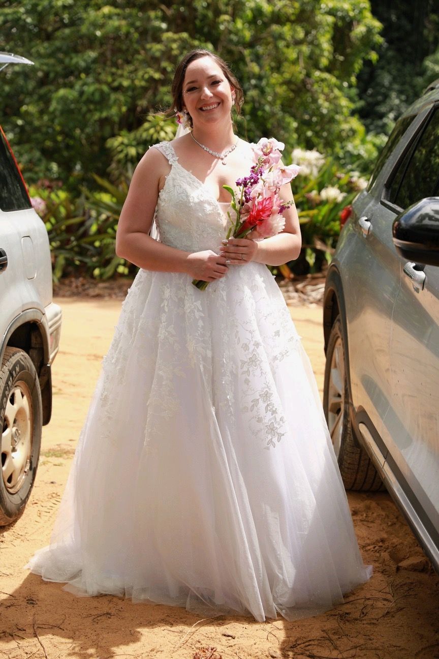 Bride in a white wedding dress holding a pink bouquet outdoors between parked cars