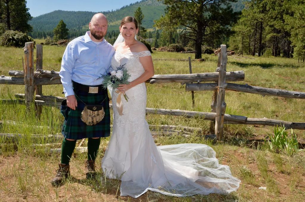 Wedding couple posing outdoors by a wooden fence, bride in white gown with bouquet and groom in a blue shirt and kilt.