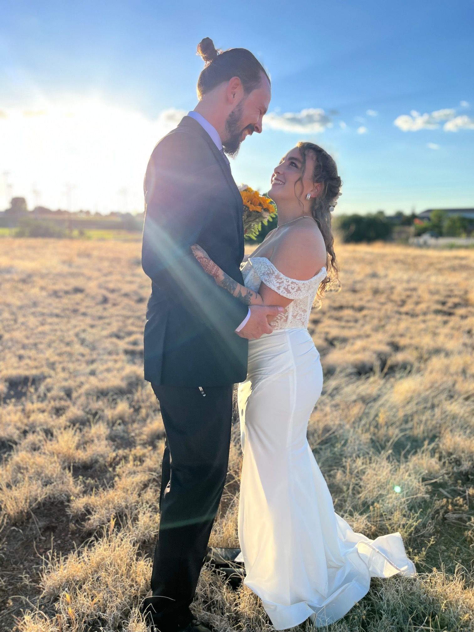 Couple embracing in a sunlit field, bride in white dress and groom in black suit at sunset