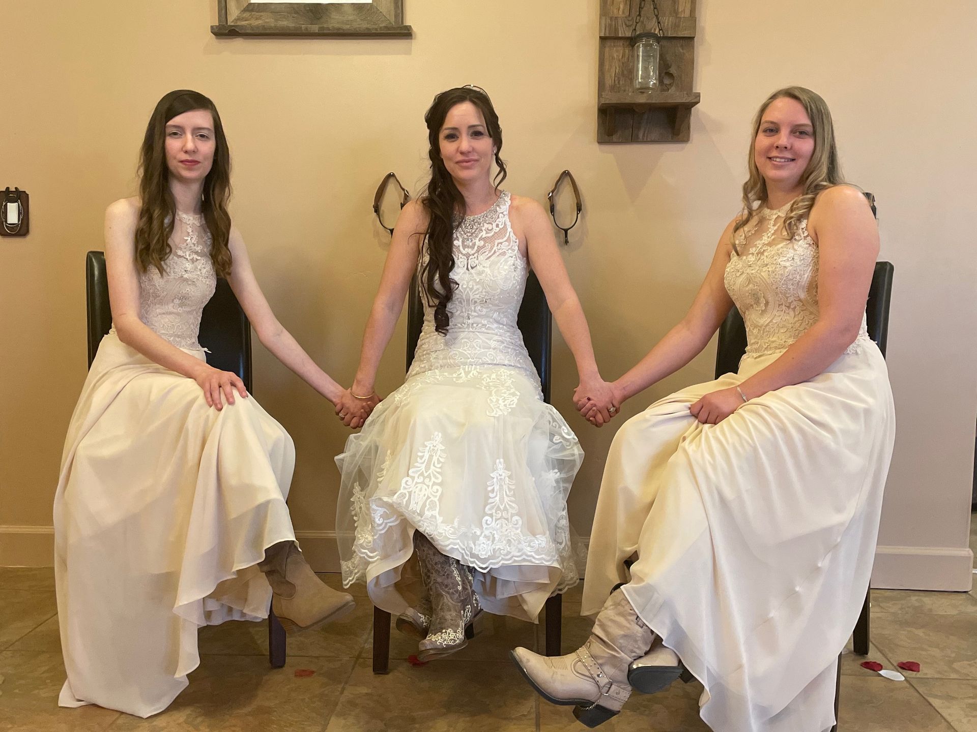 Bride and bridesmaids holding hands, seated in a room. All wearing long, light-colored dresses.