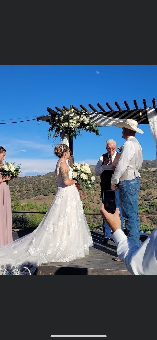 Wedding ceremony outdoors with bride in dress, groom in cowboy hat, under a floral arbor.