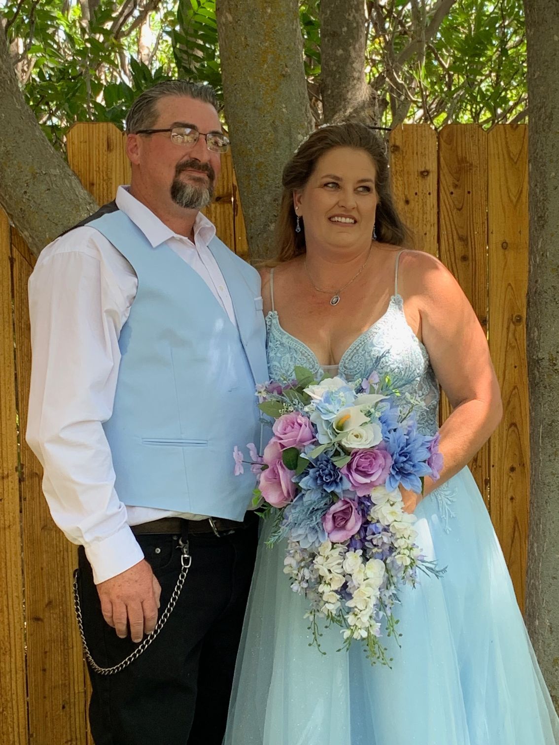 Couple in formal attire standing outdoors, holding a pastel bouquet in front of a wooden fence.
