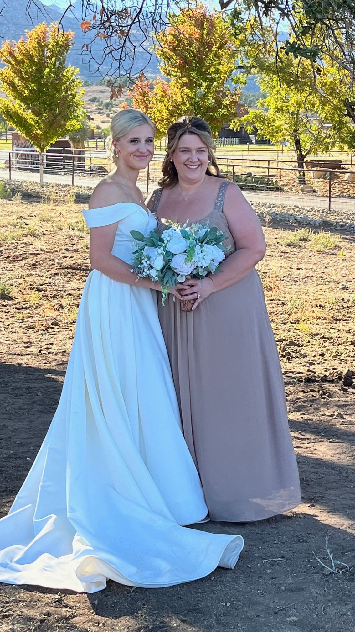 Two women in formal dresses holding a bouquet outdoors by trees and a fence.