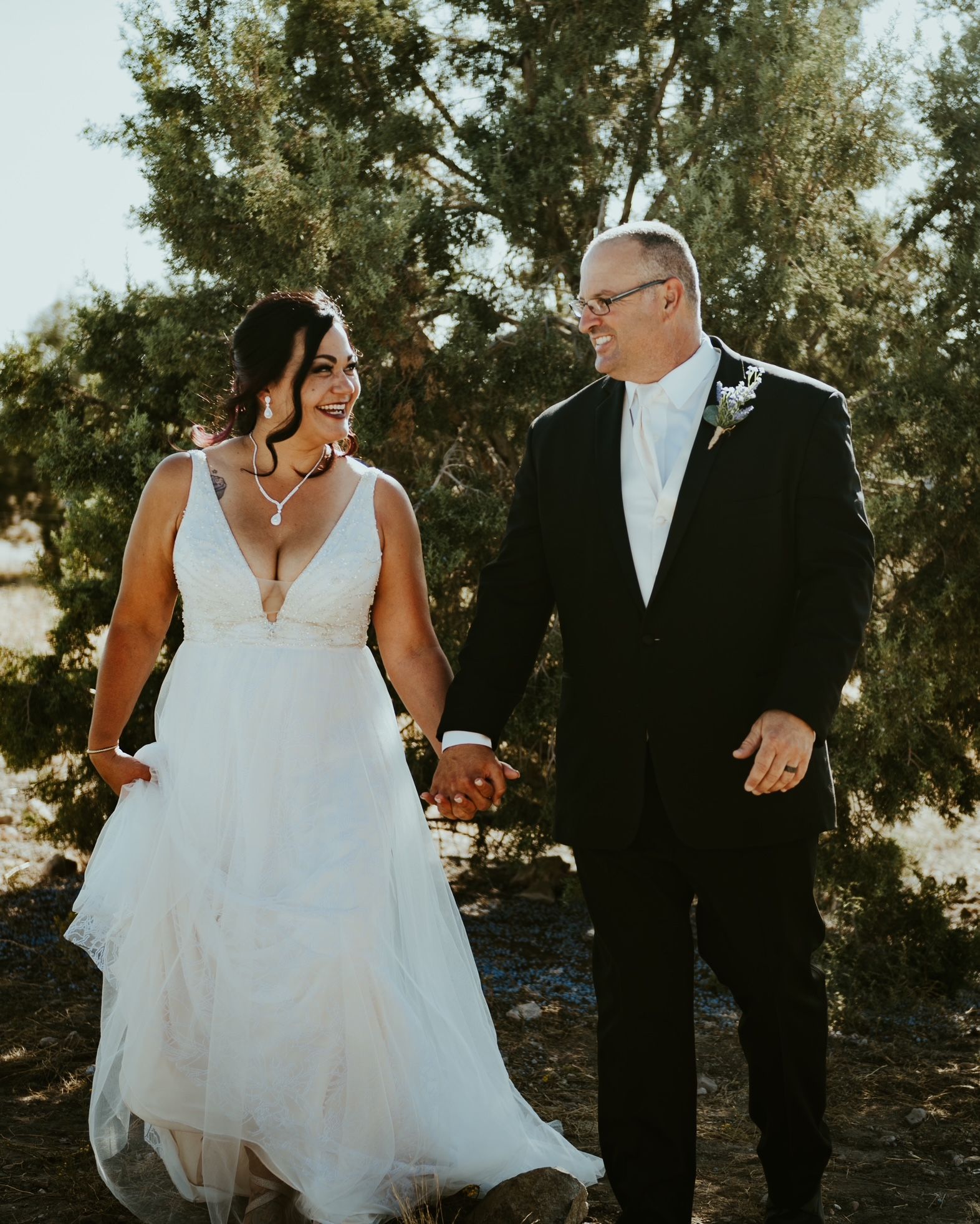 Bride and groom holding hands, smiling. Wedding attire, outdoors, sunny.