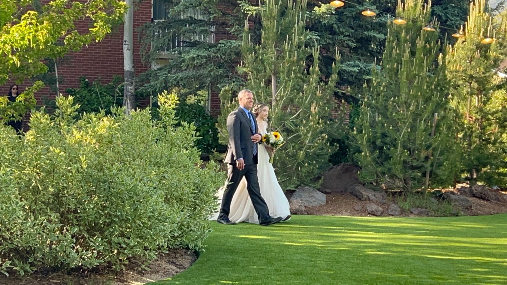 Bride and groom walk on grass after wedding ceremony, surrounded by trees and bushes.