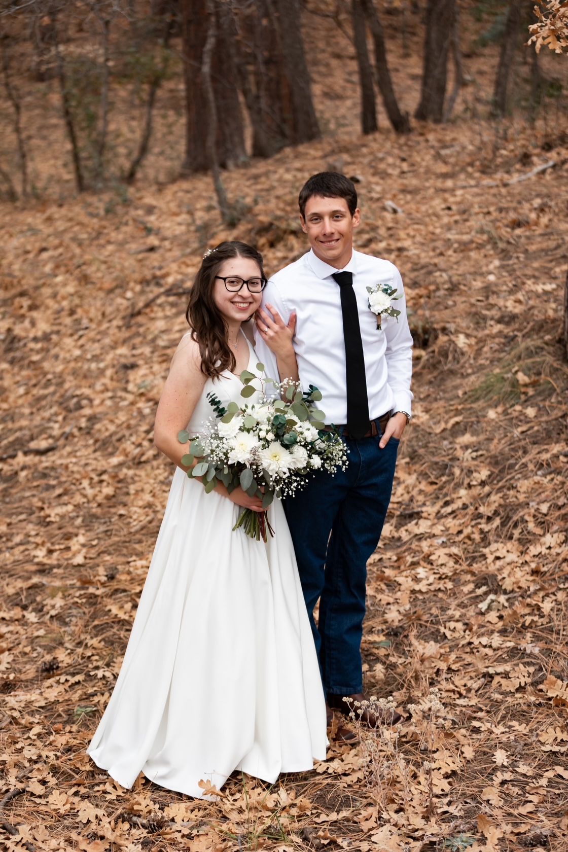 Wedding couple posing outdoors; bride holds bouquet, groom in dark tie. Fall foliage in background.