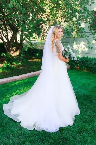 Bride in white wedding dress with a long train, holding a bouquet, standing on green grass, smiling.