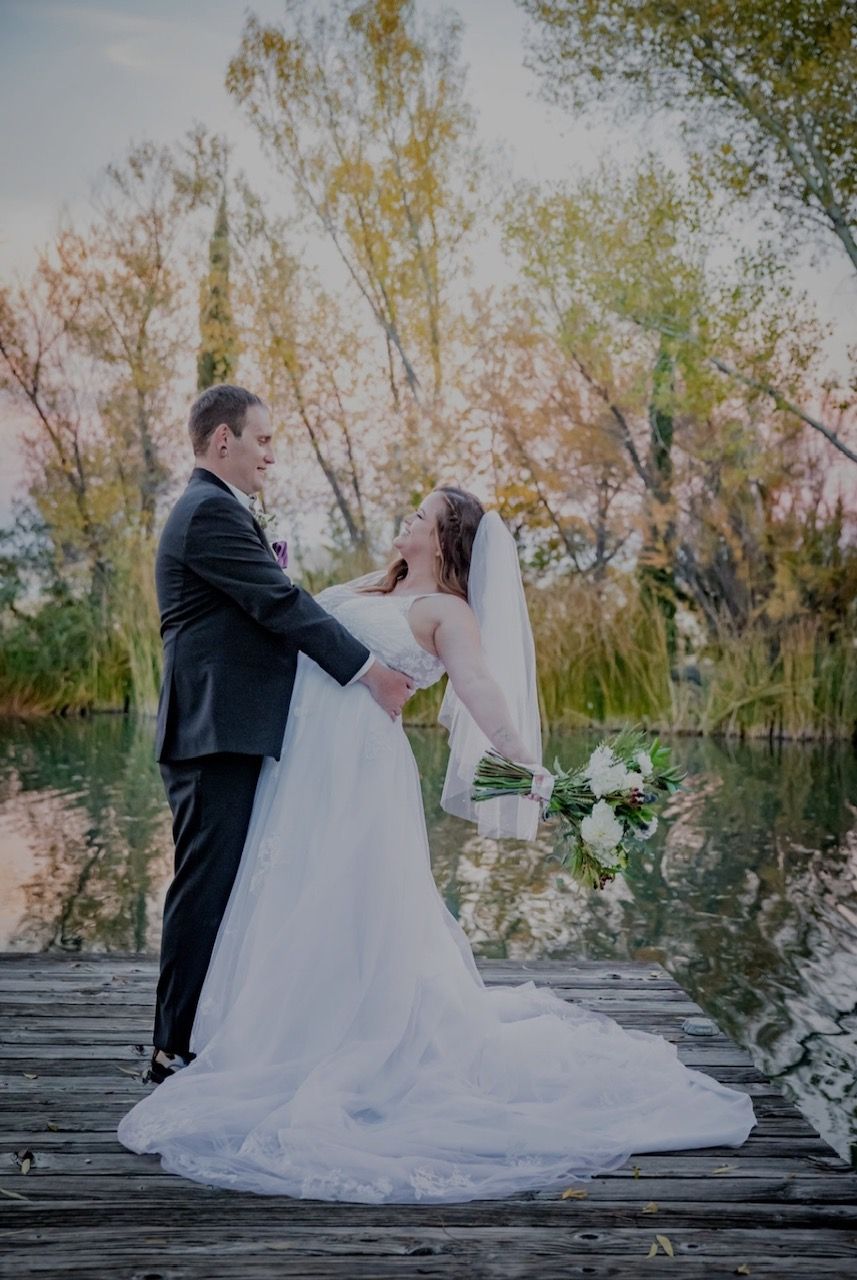 Couple dancing on a wooden dock with lake and trees in the background. Bride smiles as groom dips her.