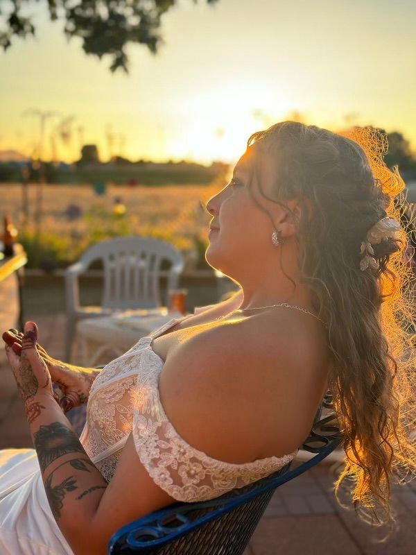 Woman in a white lace dress sits outdoors at sunset, holding a drink and facing the glowing horizon.