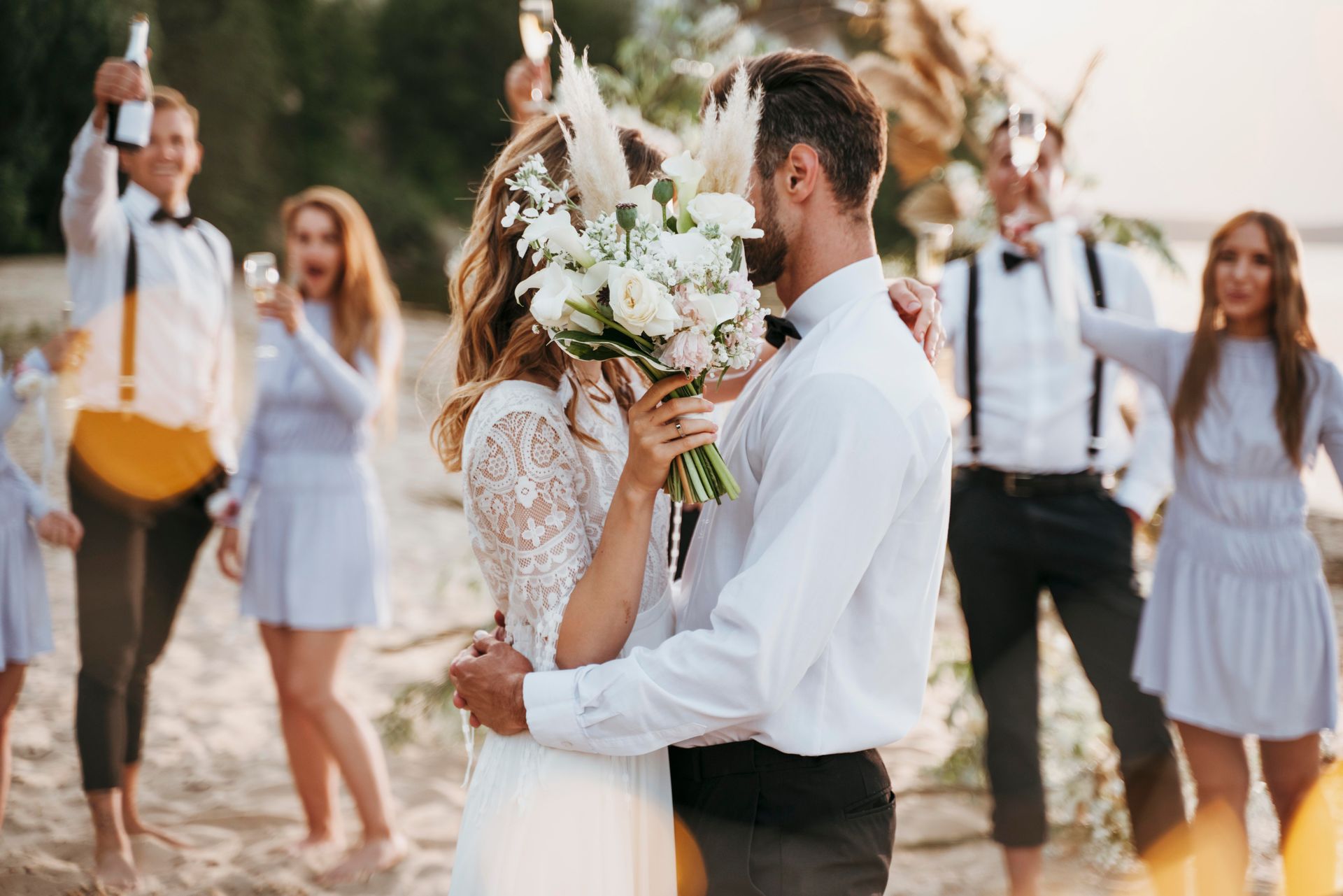 Newlyweds embrace on a beach, bouquet covers the bride's face; wedding party cheers, holding champagne, during sunset.