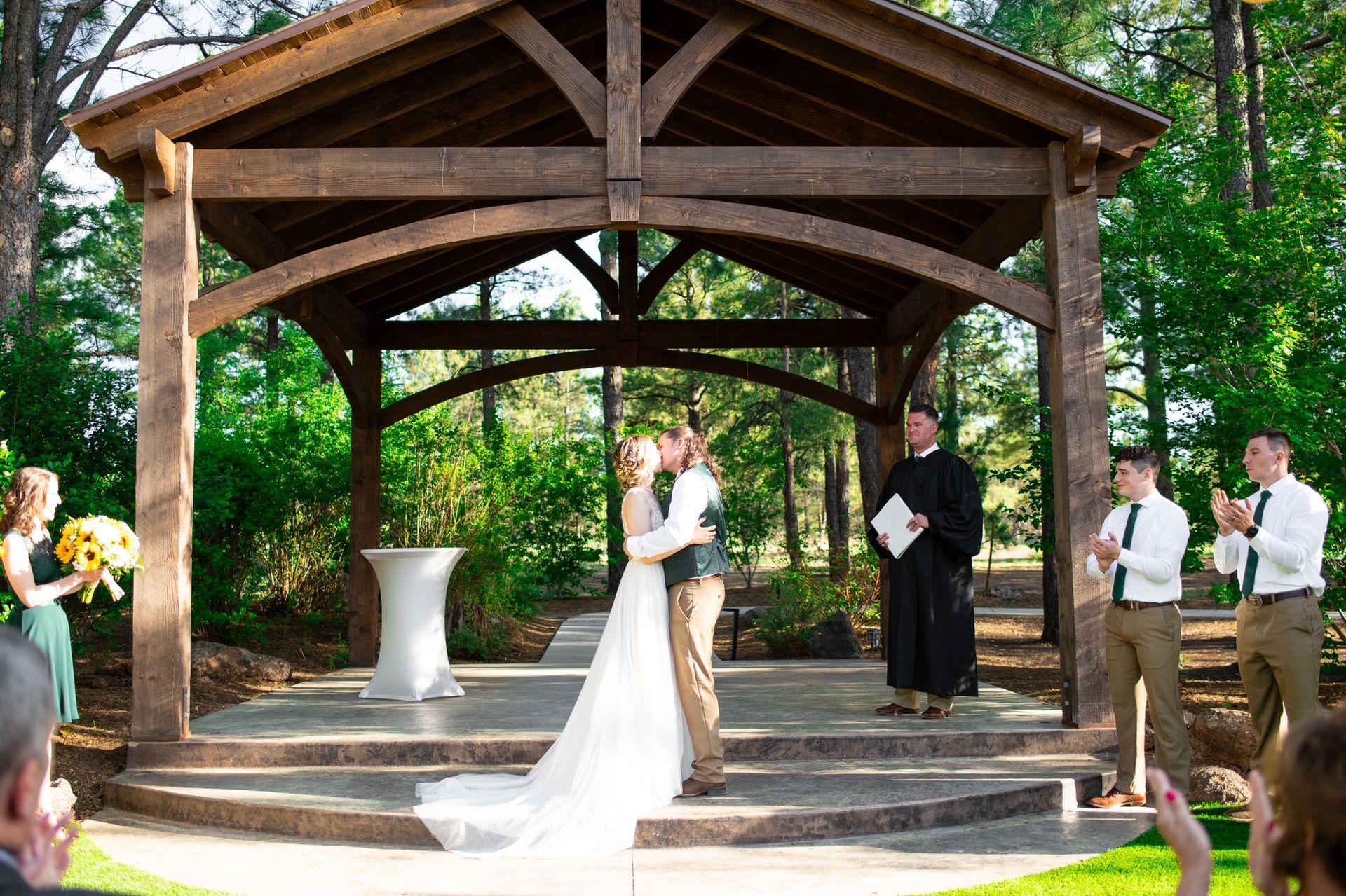 Wedding couple exchanging vows under a wooden pavilion outdoors, with officiant and guests nearby