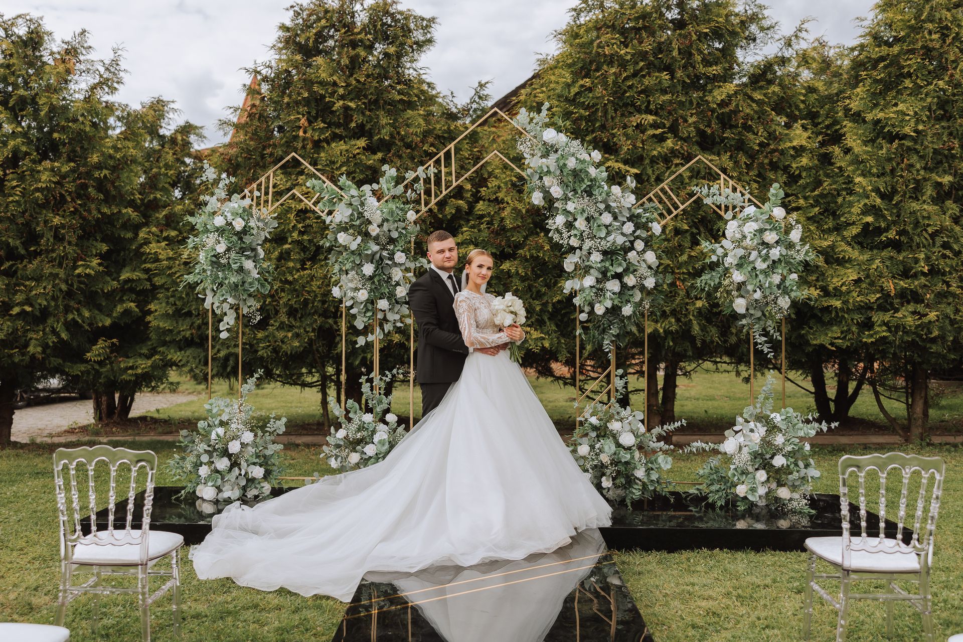 Couple embraces at wedding ceremony with floral arch and clear chairs.