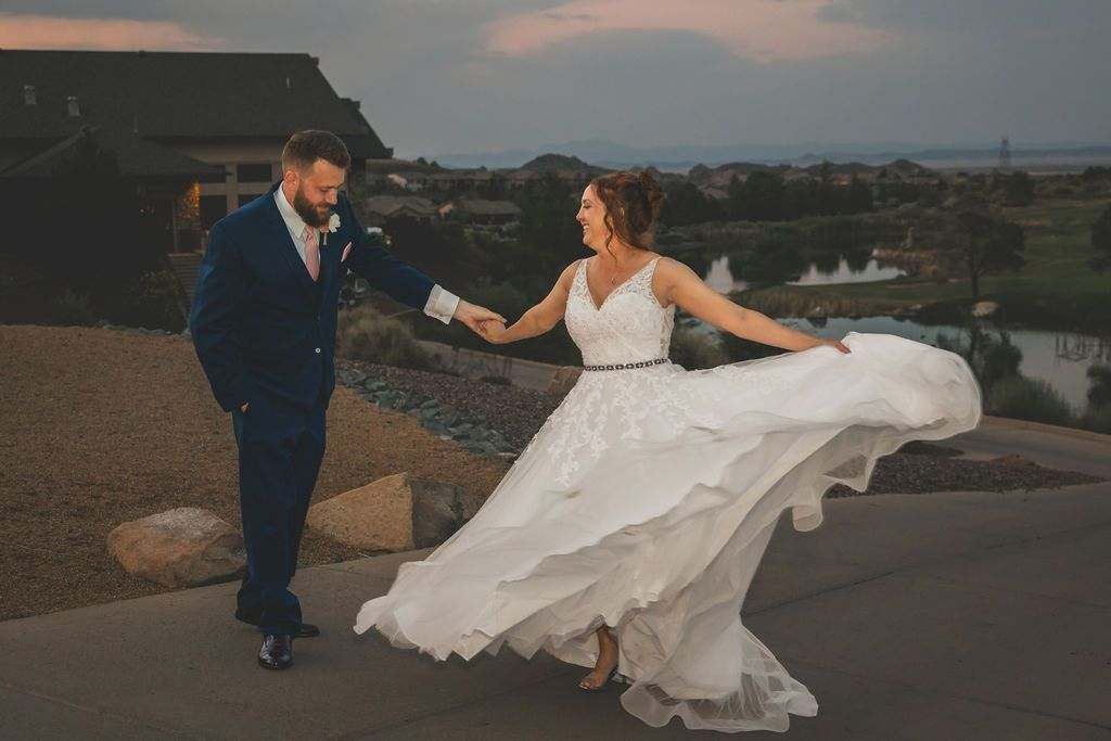 Bride in a white wedding dress twirls beside groom in a blue suit at sunset by the water