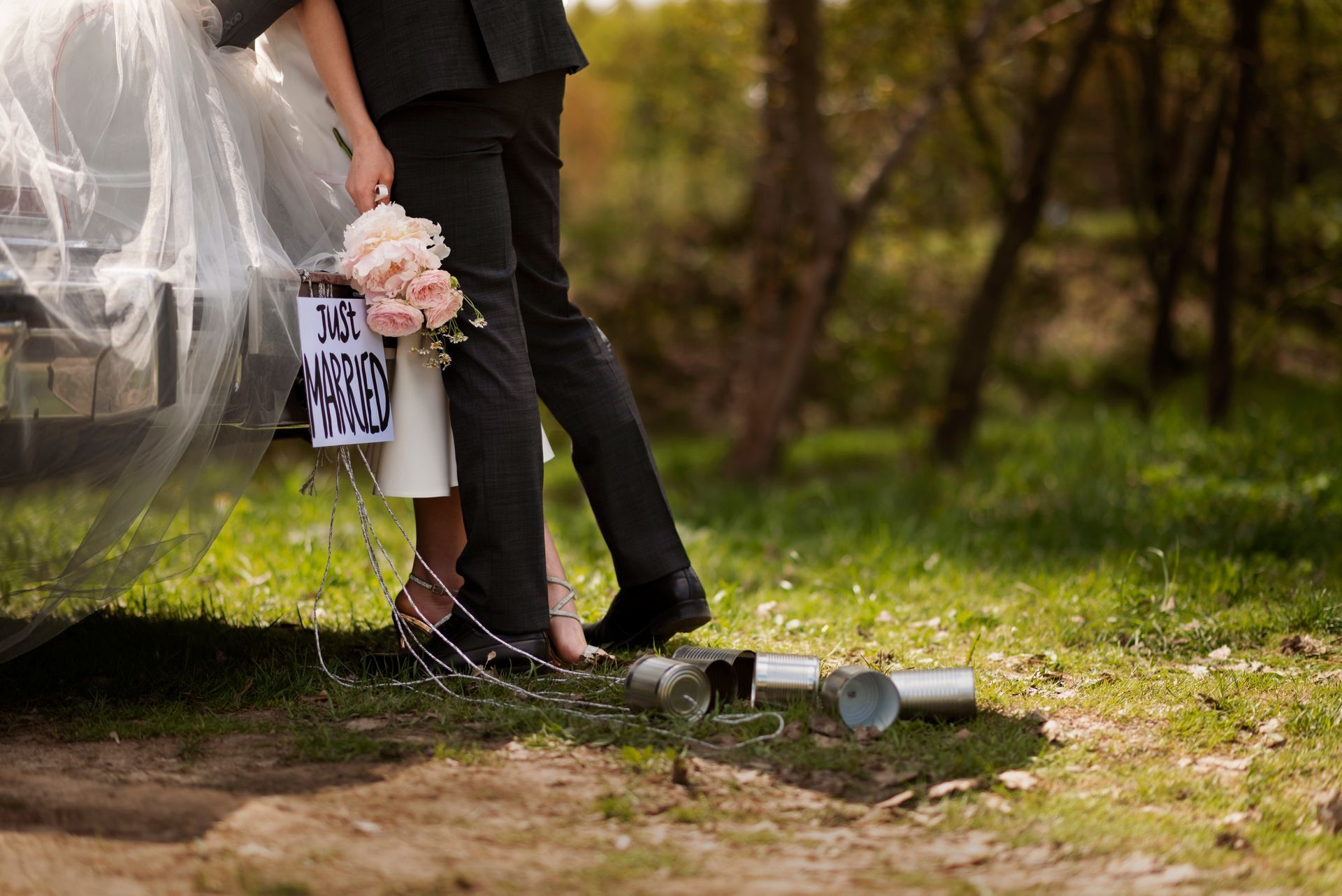 Bride and groom by a car decorated with tin cans, holding flowers.  Green grass and trees in background.