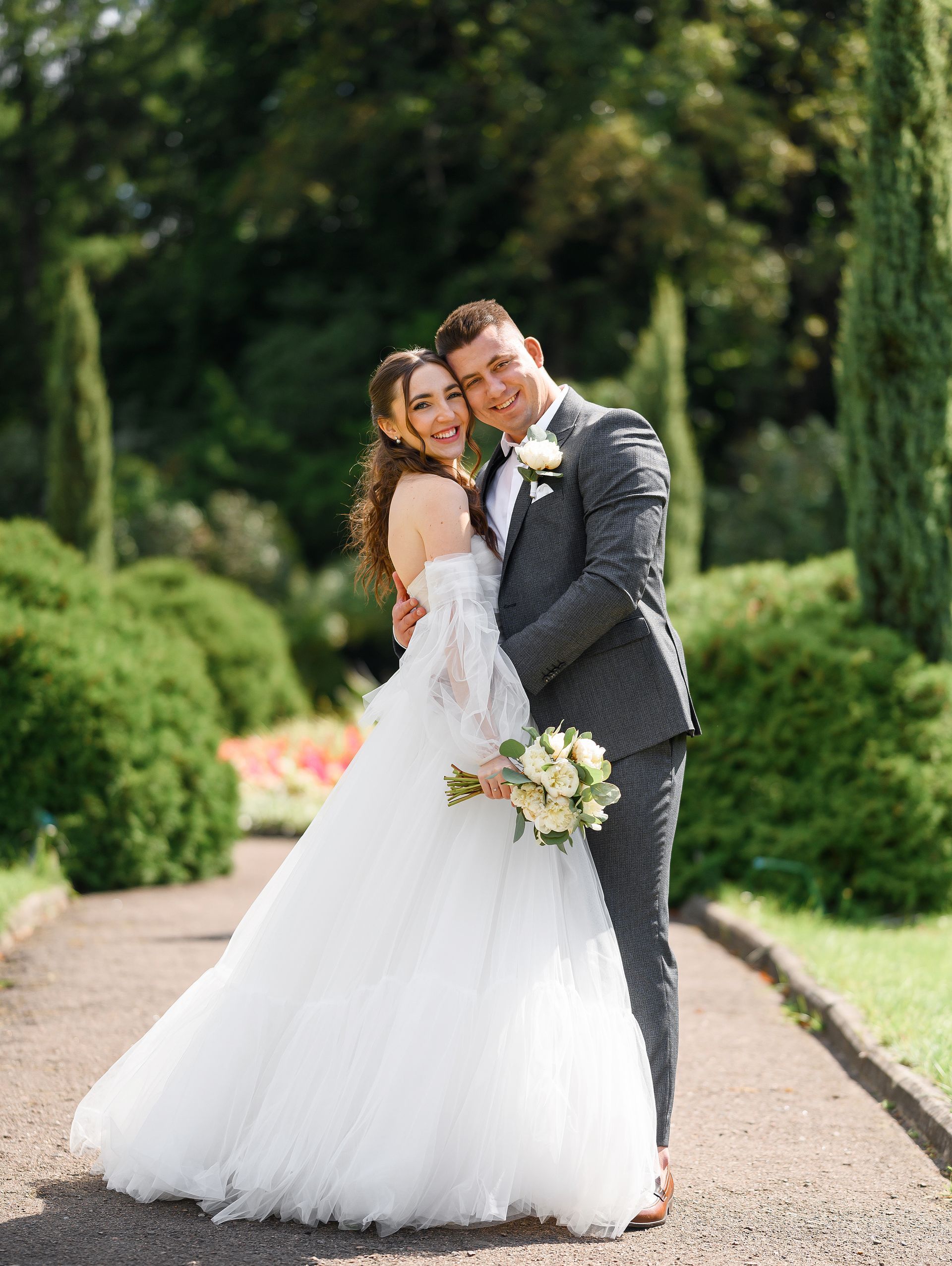 Bride and groom embrace on a garden path. She wears a white gown, he wears a gray suit, both smile.