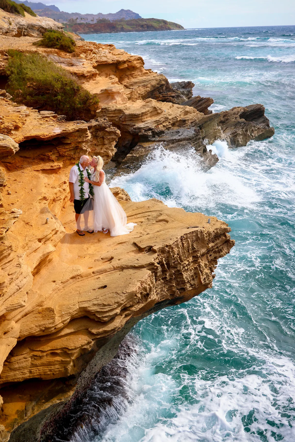 Couple in wedding attire on a rocky cliff overlooking ocean with crashing waves.