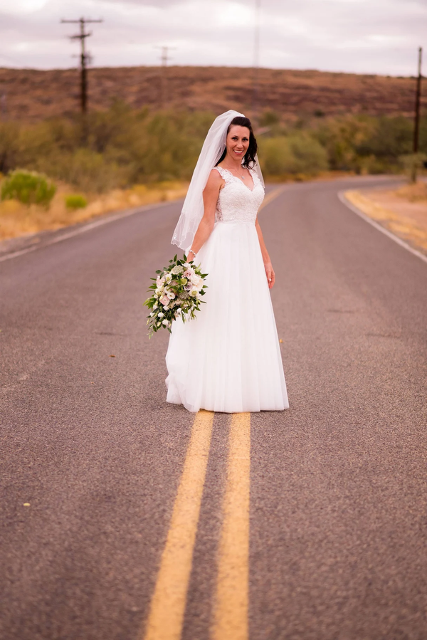 Bride in white dress holding flowers, standing on a road with yellow lines, in a rural landscape.