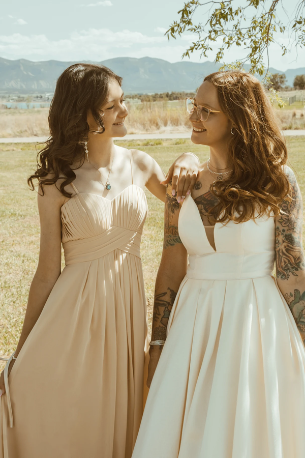 Two women smiling, dressed in elegant gowns, outdoors. One has tattoos; mountain backdrop.