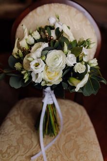 Bouquet of white flowers with green foliage, tied with a white ribbon, resting on a chair.