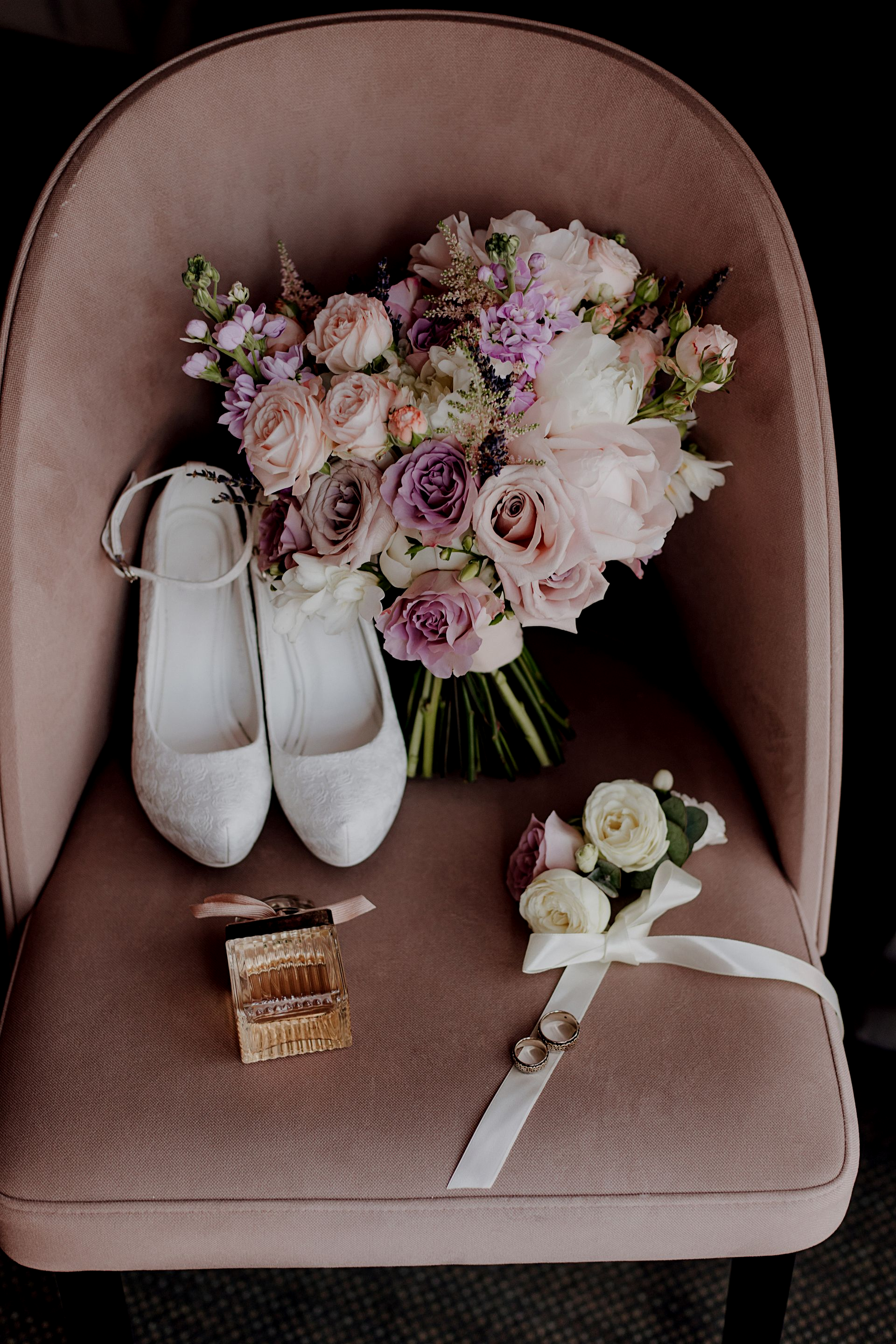 Wedding details: bouquet, shoes, boutonniere, and rings on a pink chair.