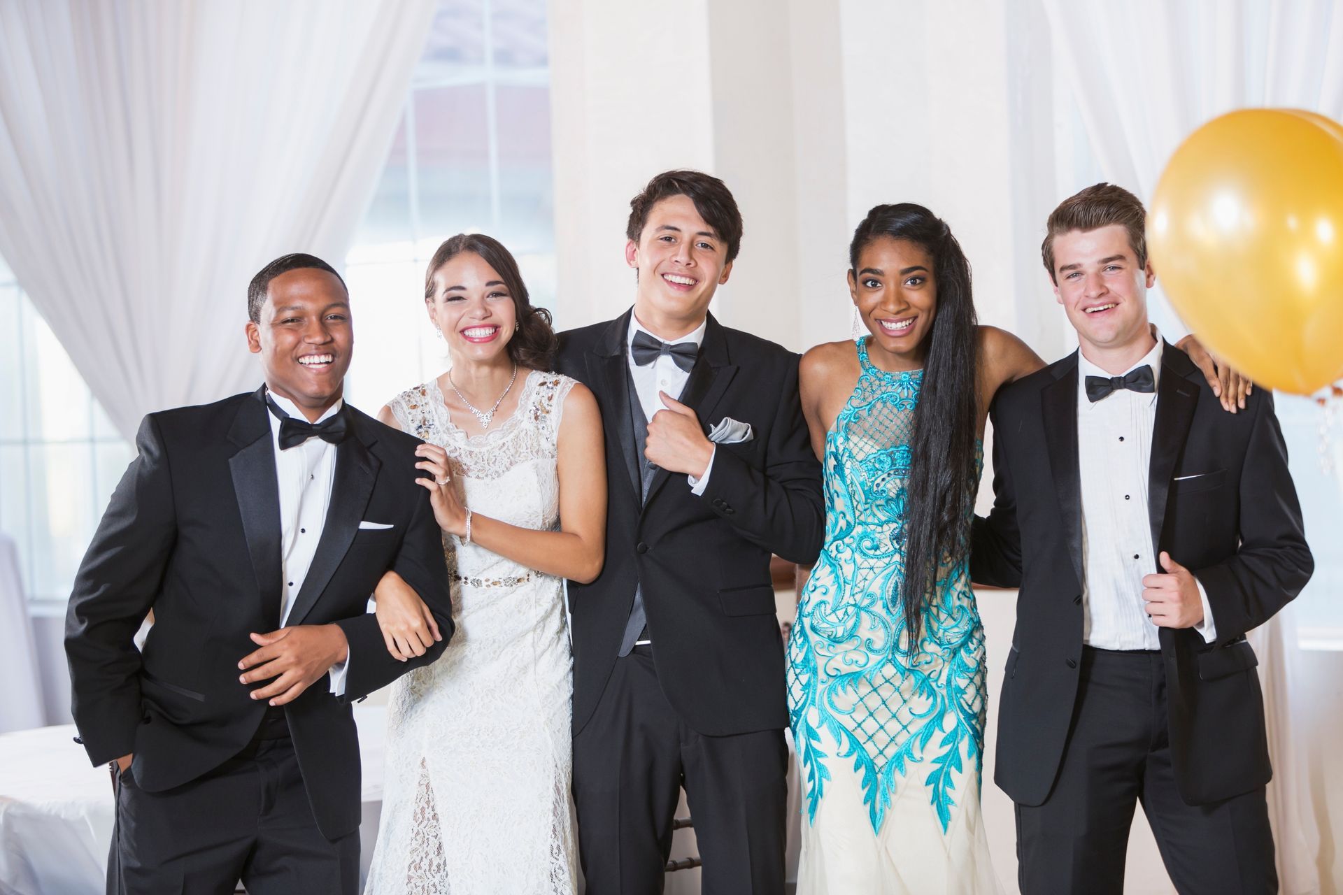 Five people in formal attire pose for a photo. One holds a gold balloon. They smile and stand near a window.