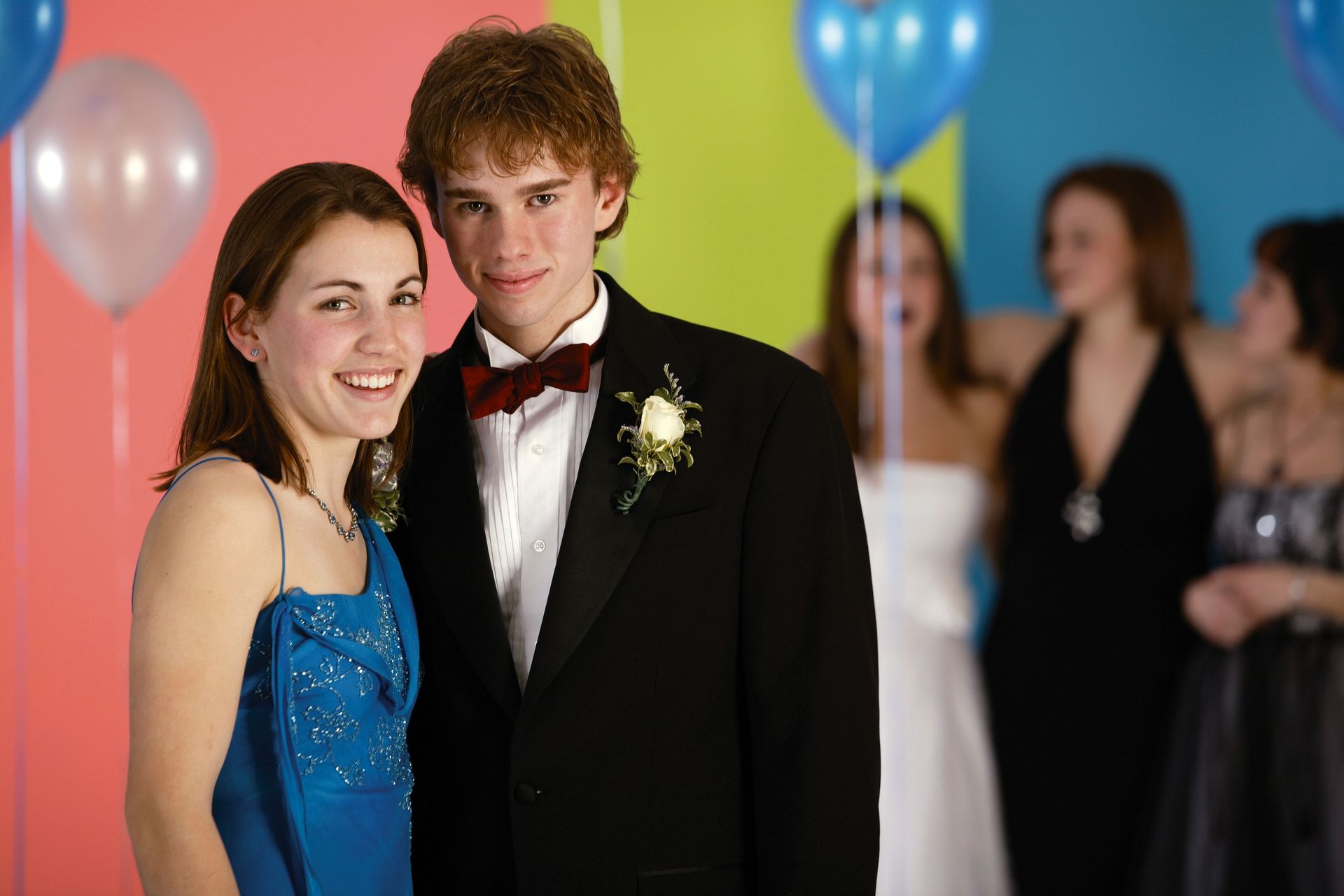 Couple at prom, she in blue dress, he in tuxedo, balloons in background, others blurred.
