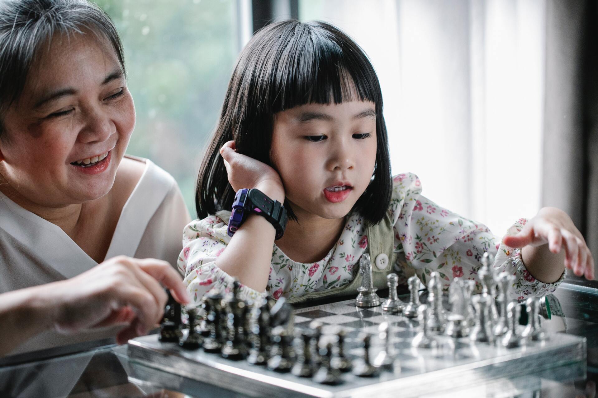 Woman smiles, teaching girl chess. Both hands on metal chessboard in a well-lit room.