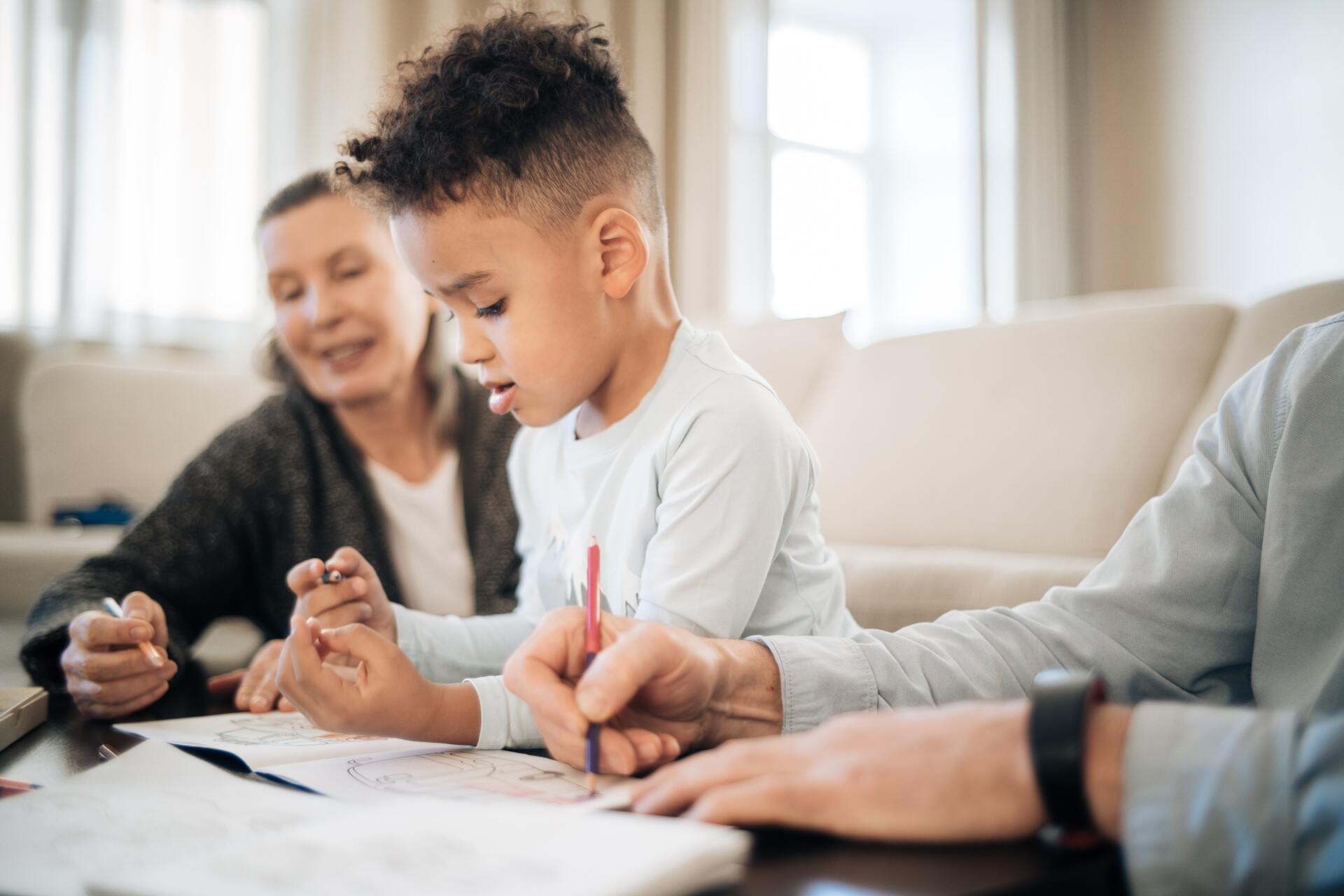 Boy and adults sitting at table, drawing together; light-colored room.