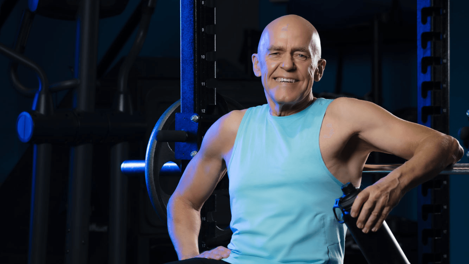 Bald man in blue tank top smiles, leaning on weight machine in a gym.