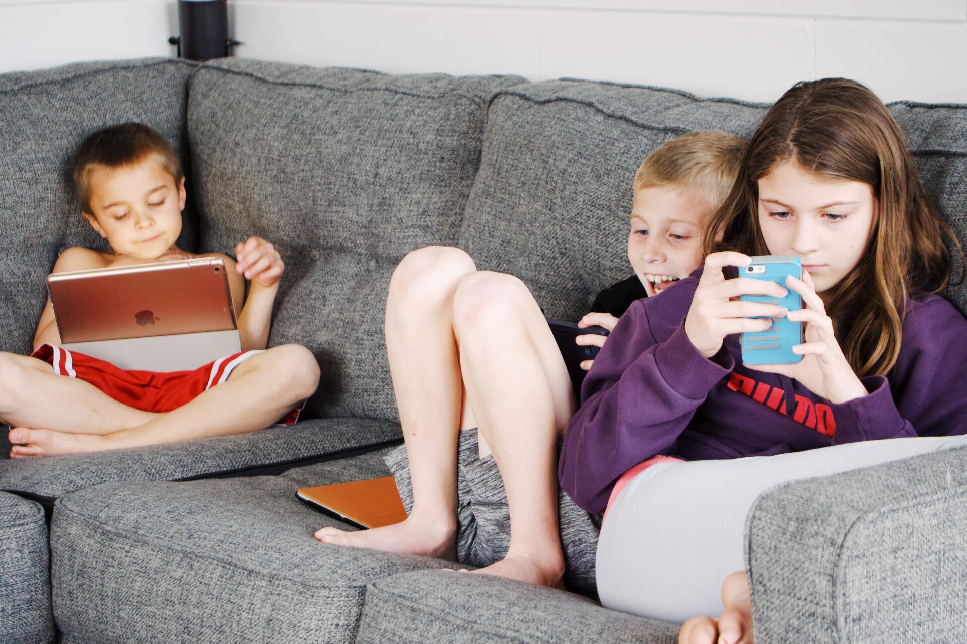 Three children on a gray couch, each using a digital device: tablet, phone, and another tablet.