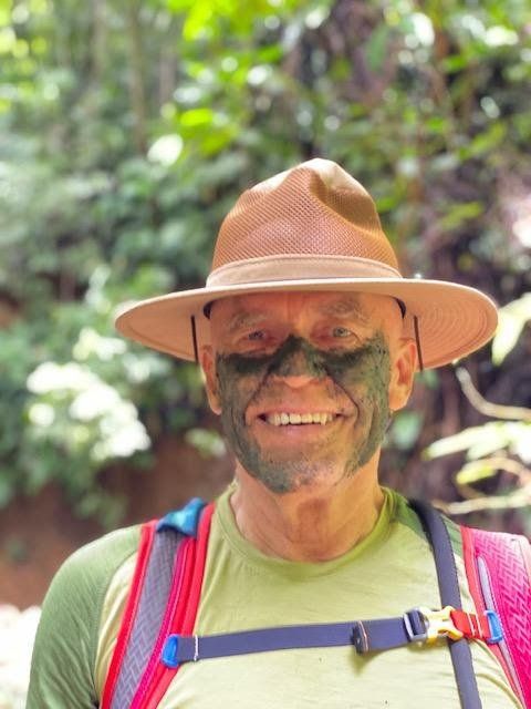 Man in brown hat and camouflage face paint smiling outdoors.