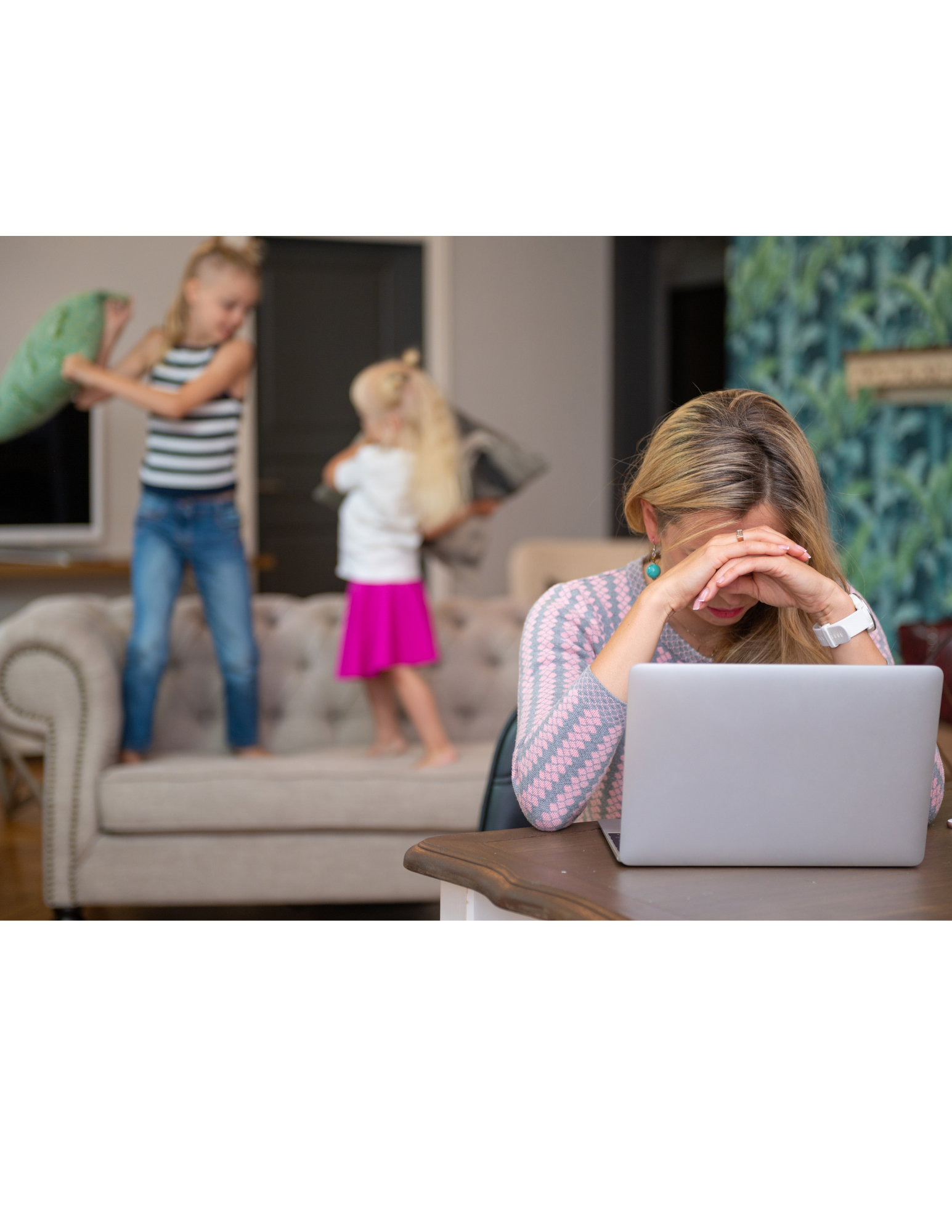 Woman with hands on face, looking stressed at laptop. Two children play-fight on couch in background.
