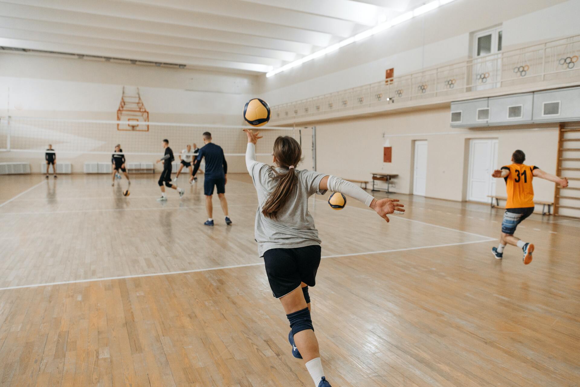 People playing volleyball in a gymnasium. A player in a grey shirt prepares to serve the ball.