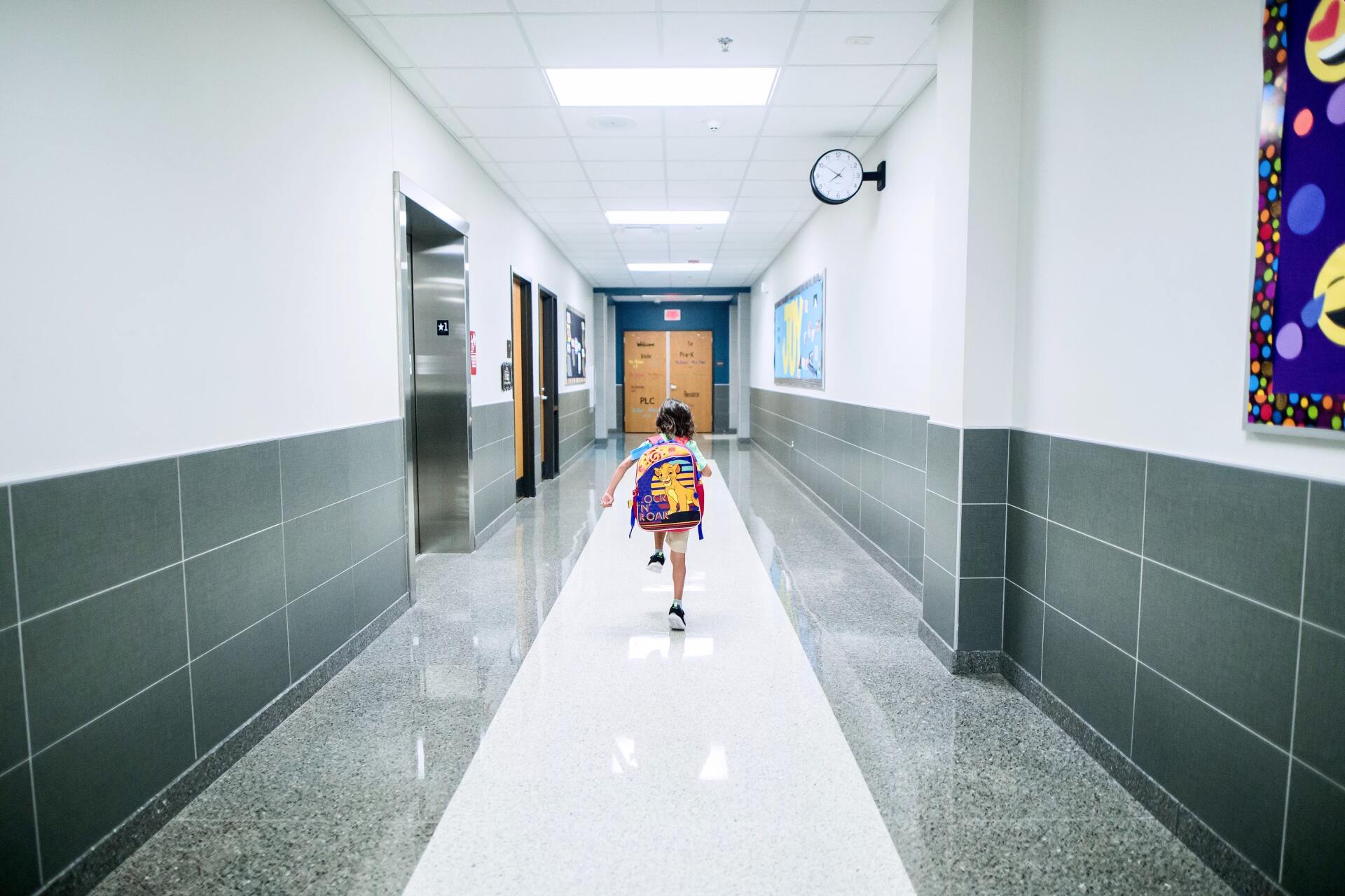 Child with backpack running down a school hallway towards a set of doors.