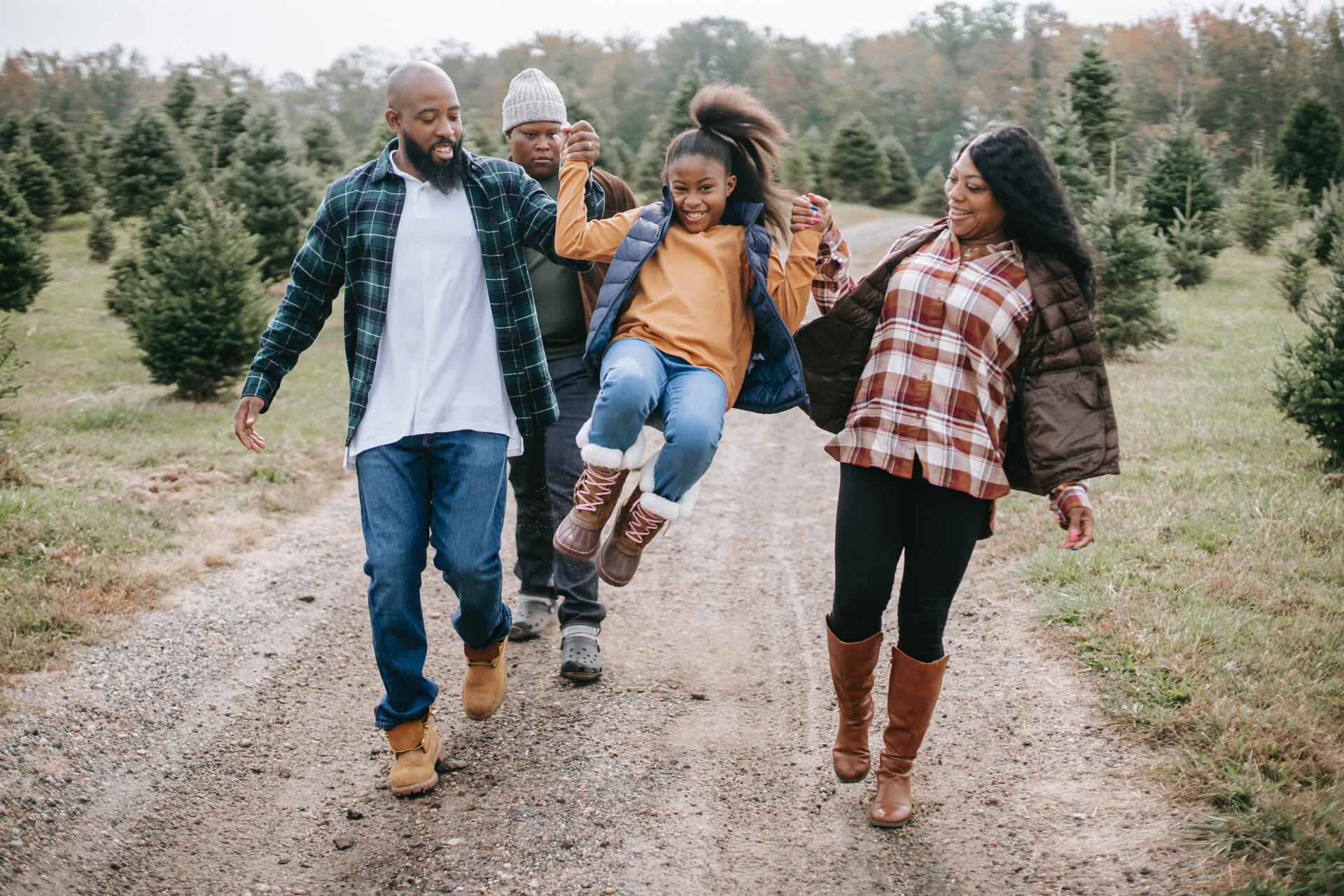 Family of four smiling, laughing, walking down dirt path in Christmas tree farm.