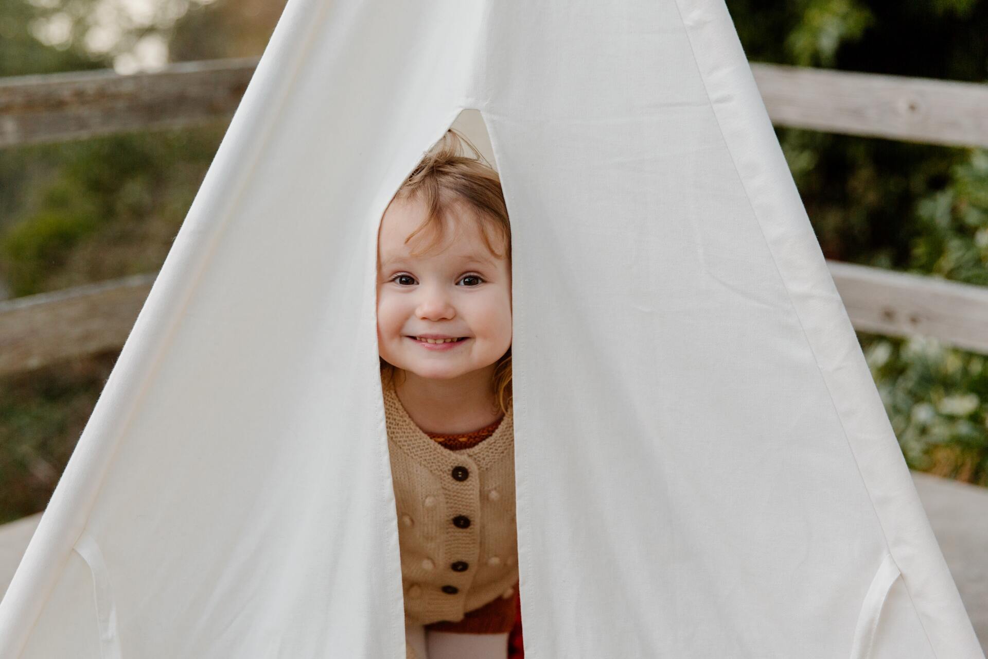 Smiling child peering out from inside a white teepee outdoors.