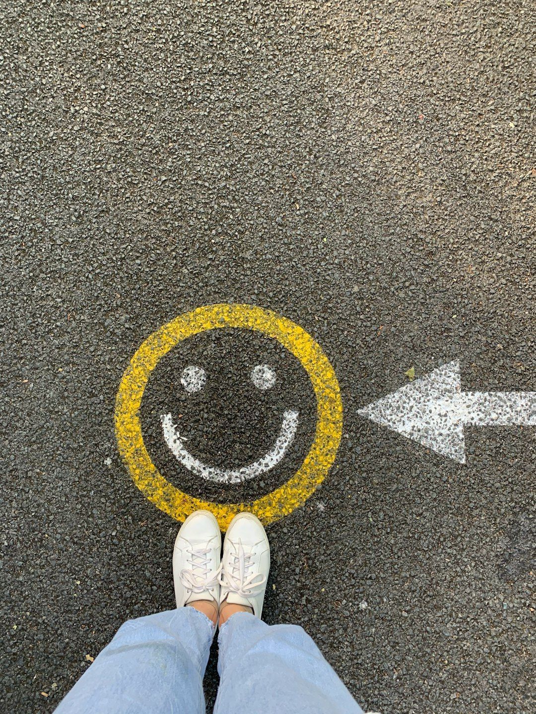 Feet wearing white sneakers standing in front of a yellow smiley face on pavement, an arrow points right.