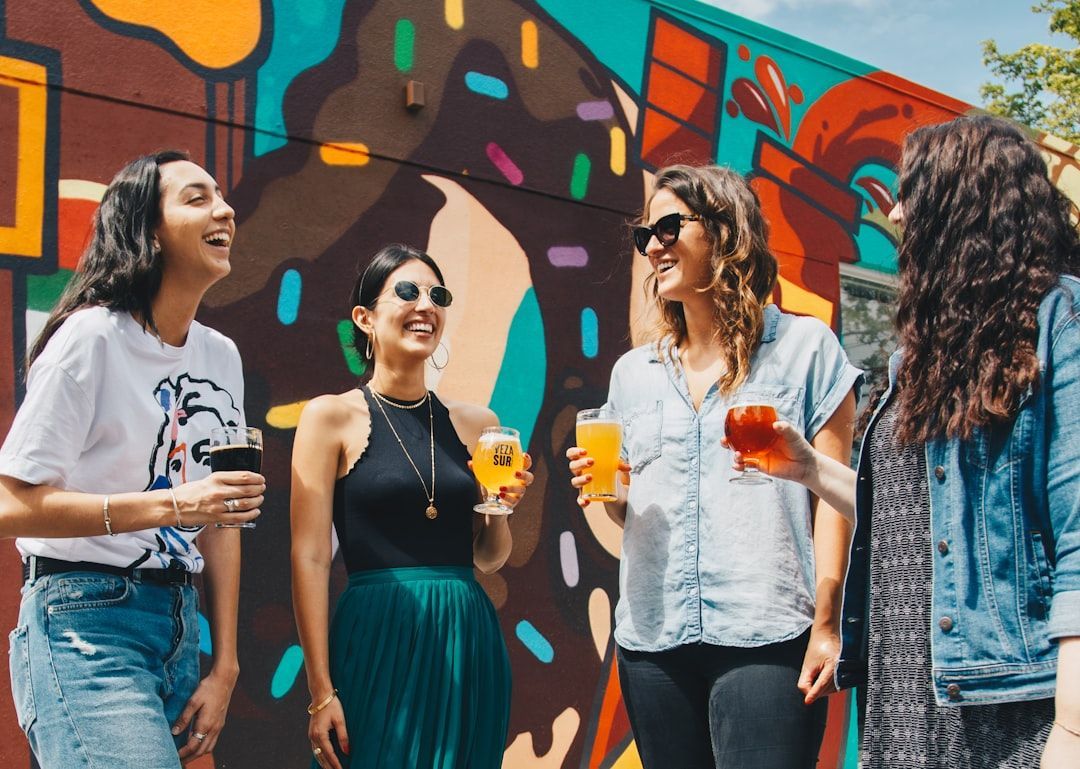 Four women laughing, holding drinks, in front of a colorful mural on a sunny day.
