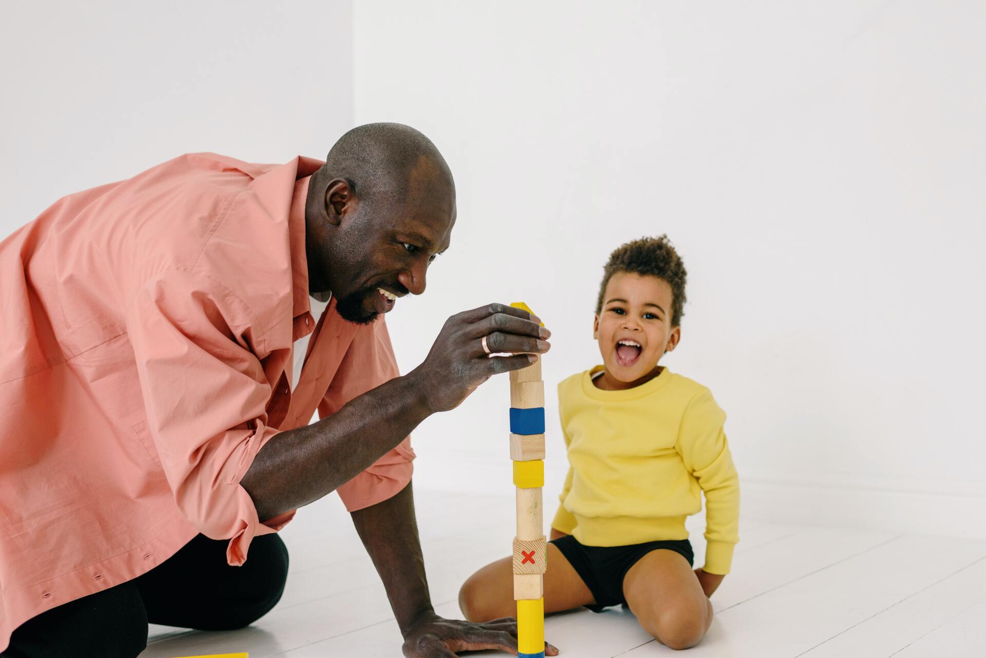 Man and child building a tower of colorful wooden blocks on a white floor. Child is smiling.